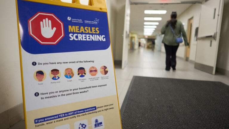 Canada just lost its measles elimination status. Is the U.S. next? A sign advertising measles screening is in the foreground, to the left. The top of the sign has a red octagon with a white hand signaling stop, with "measles screening" written to the right. Underneath the sign lists measles symptoms and what to do if exposed to measles. To the right of a sign, a woman walks through open double doors into a hospital corridor.